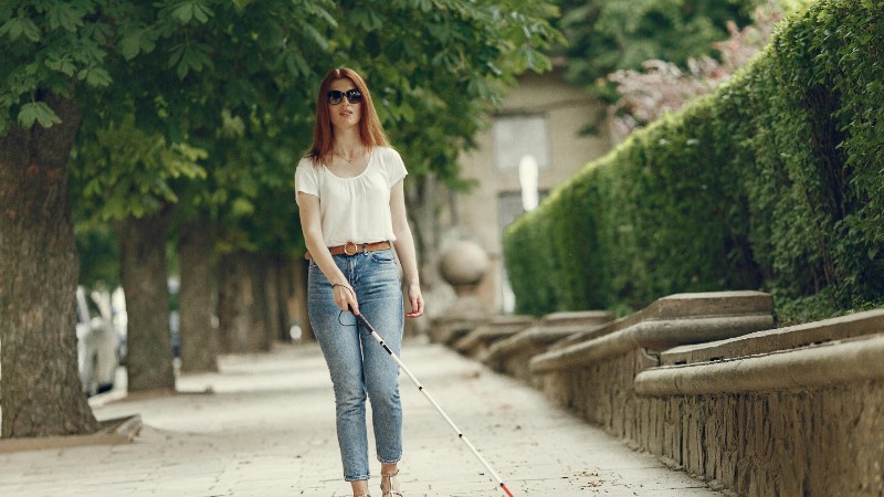 A visually impaired woman walks confidently using a cane on a tree-lined urban sidewalk., tags: bastone intelligente - pexel